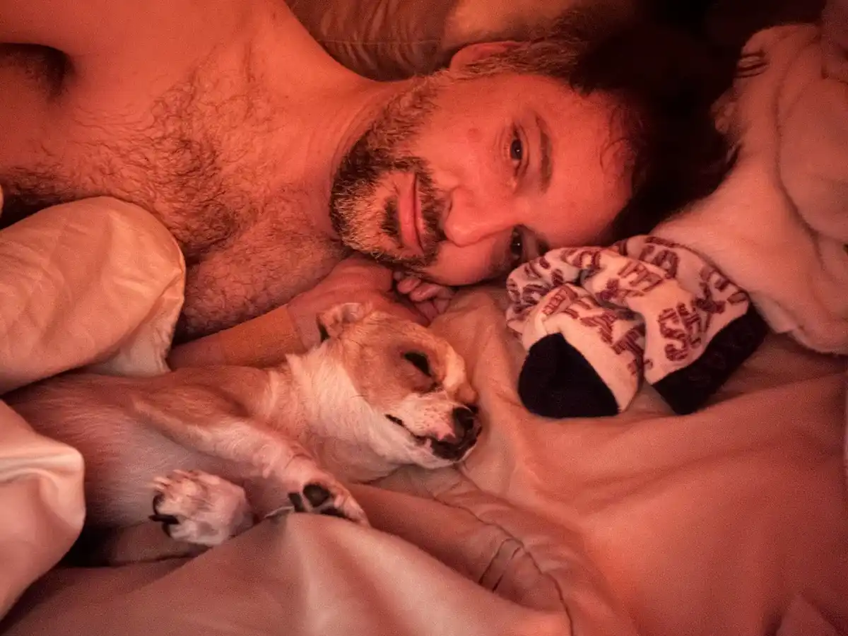 A shirtless man with a beard lies in bed under warm lighting, looking at the camera while a small dog sleeps peacefully…
