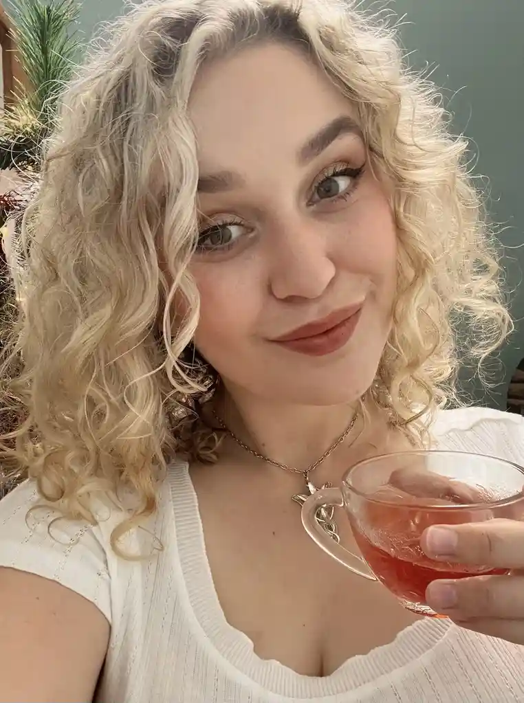 A person with curly blonde hair and a white top holds a glass cup of pink liquid, looking at the camera with a slight smile…