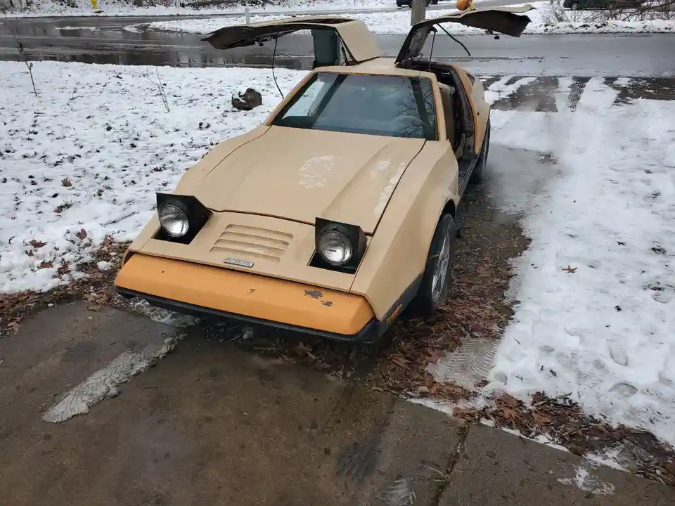 A beige and orange vintage sports car with gull-wing doors open, parked on a snowy driveway with melting snow and fallen…