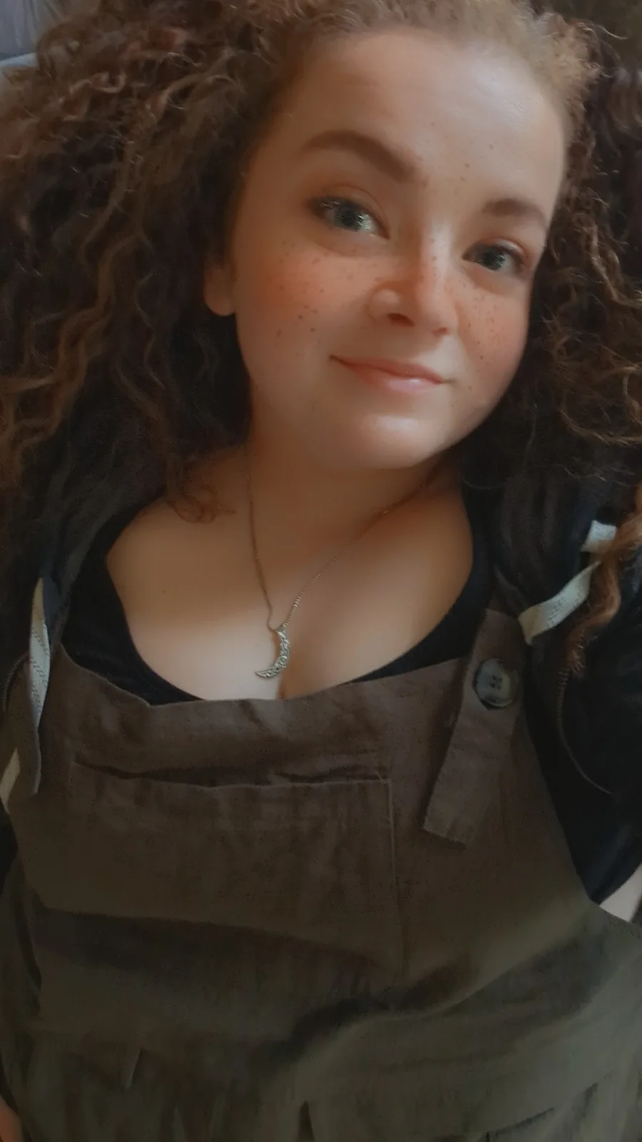 A young woman with curly brown hair and freckles smiles at the camera while wearing olive green overalls over a black top…