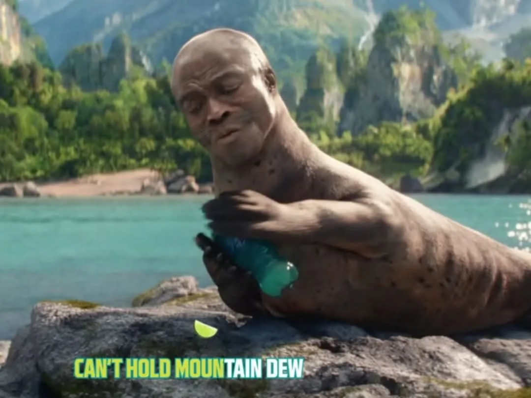 A bald, muscular man with a walrus-like face is sitting on a rock by a turquoise lake, struggling to hold a green Mountain…