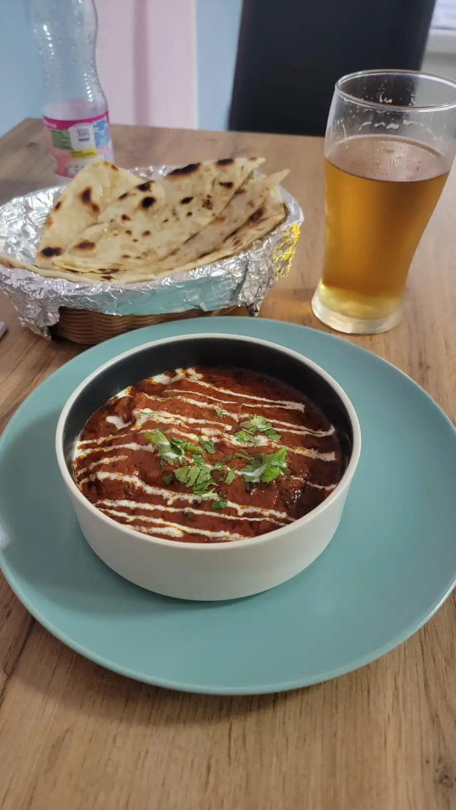 A bowl of red curry with cream drizzle and cilantro sits on a teal plate, accompanied by a basket of naan bread and a glass…