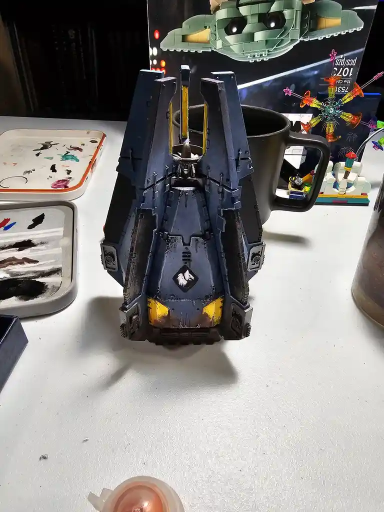 A painted miniature model, resembling a sci-fi spaceship or vehicle, is shown sitting inside a black mug on a white work…