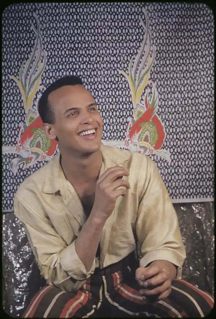 A smiling man in a vintage photo, wearing a light-colored shirt and striped pants, sits against a patterned backdrop with…