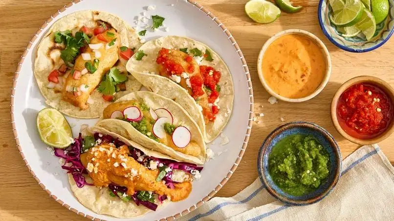 An overhead shot of three fish tacos on a white plate, each with different toppings like pico de gallo, radishes, and…