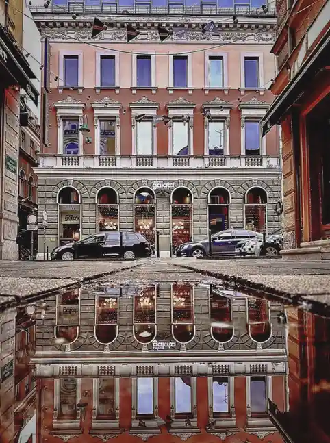 A photograph of a European-style building with ornate architecture and arched windows, reflected in a puddle on a wet…