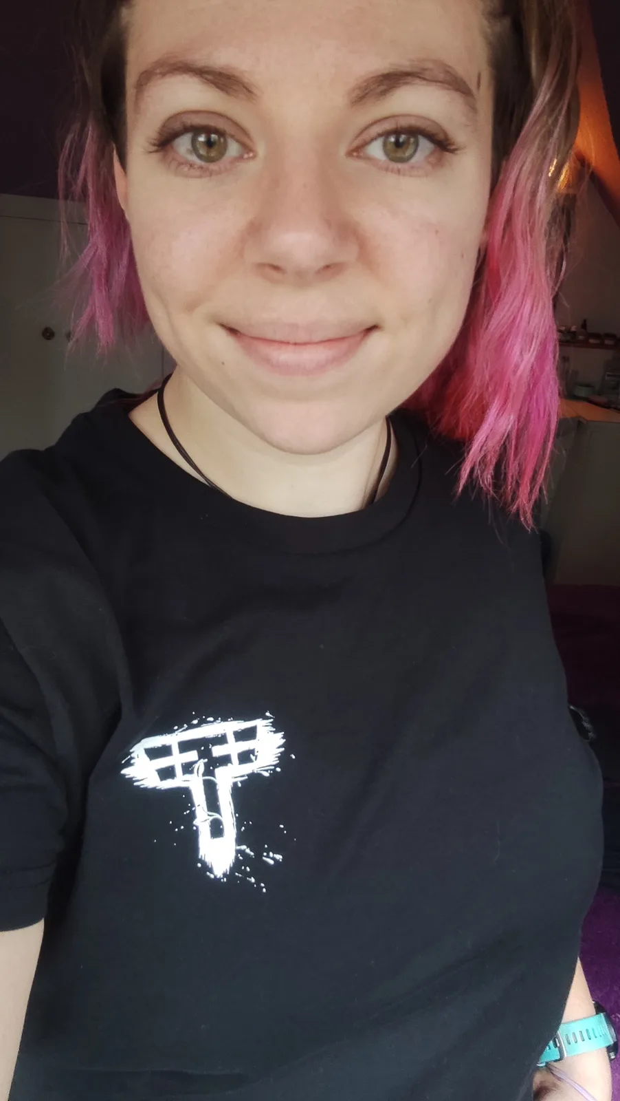 A person with pink hair and a black t-shirt featuring a white, stylized Tesla logo smiles at the camera. The overlaid text…