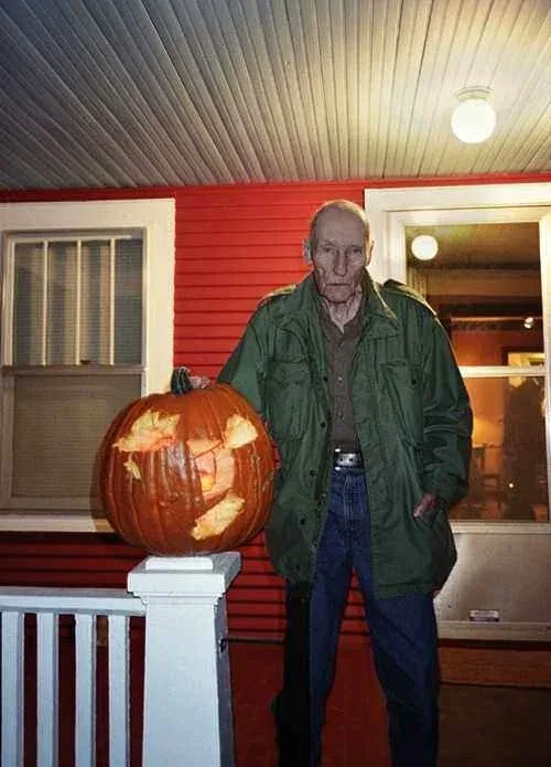 An elderly man in a green jacket stands on a porch next to a large, carved pumpkin resting on a white post. The pumpkin has…
