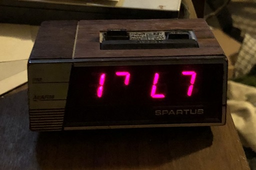 A vintage wooden digital alarm clock with red LED display shows '17 L 7' instead of a normal time. The brand 'SPARTUS' is…