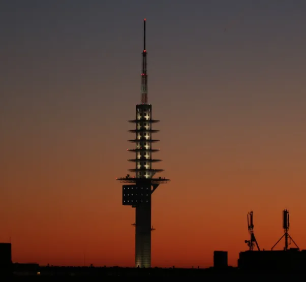 A tall, illuminated tower with a distinctive tiered structure stands against a sunset sky, its lights glowing against the…