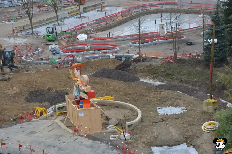 An aerial view of a construction site with dirt, machinery, and red safety barriers. A large, colorful statue of Woody from…