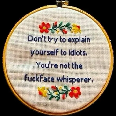 A circular embroidery hoop holds a piece of white fabric with blue stitched text that reads 'Don't try to explain yourself…