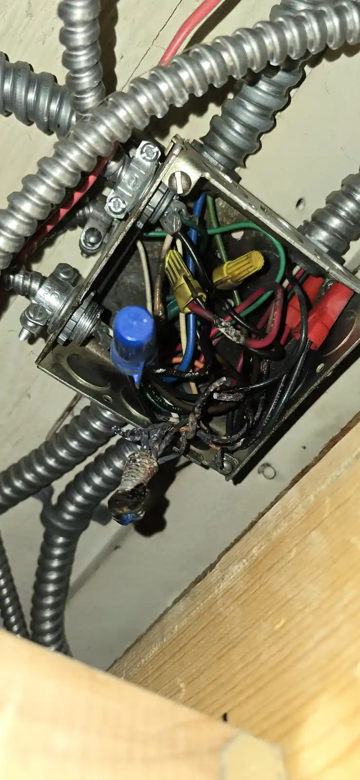 A close-up photo shows a messy, exposed electrical junction box with tangled wires of various colors, some with burnt…