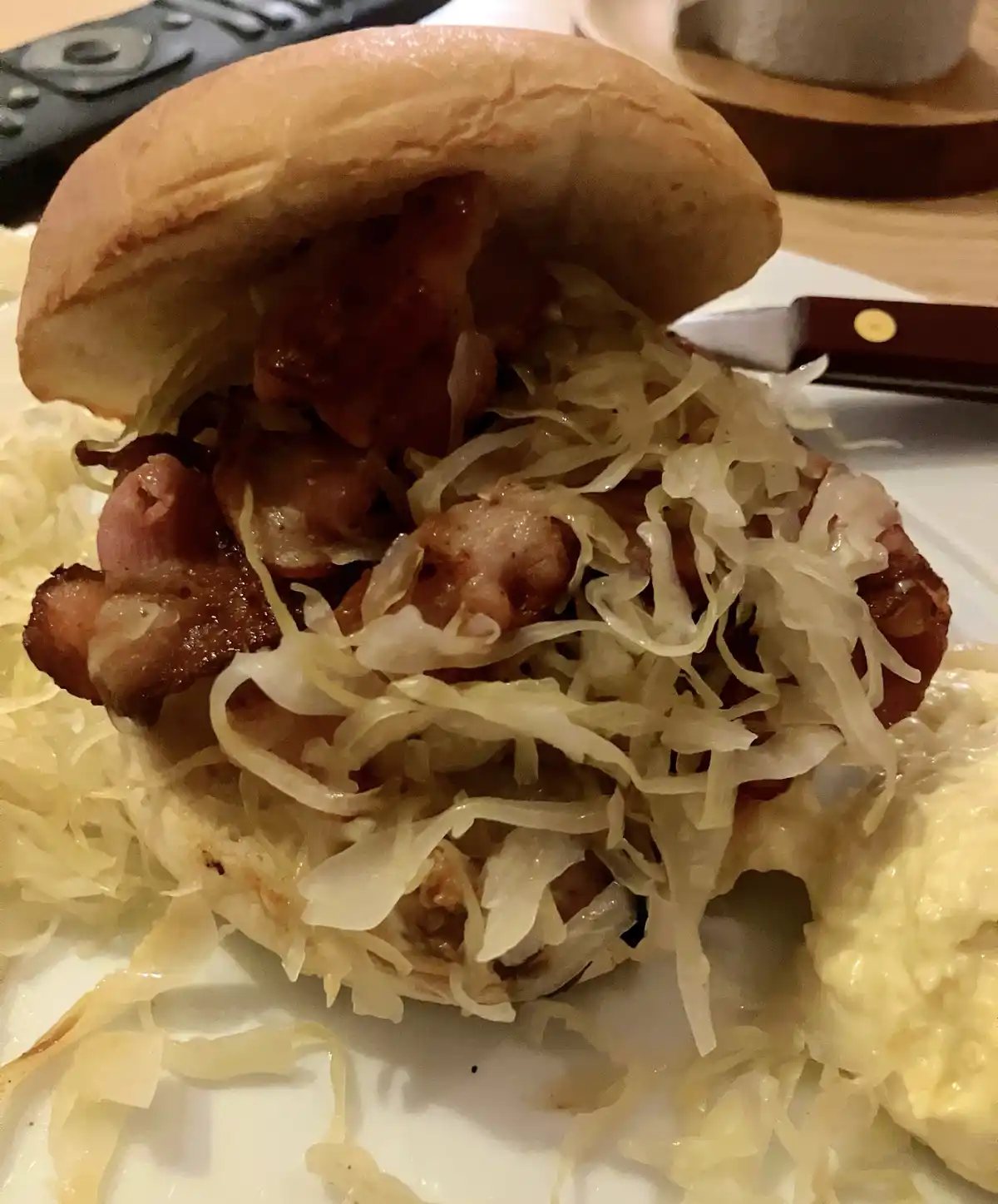 A close-up photo shows a sandwich on a white plate, filled with sauerkraut and pieces of bacon, with a knife and remote…
