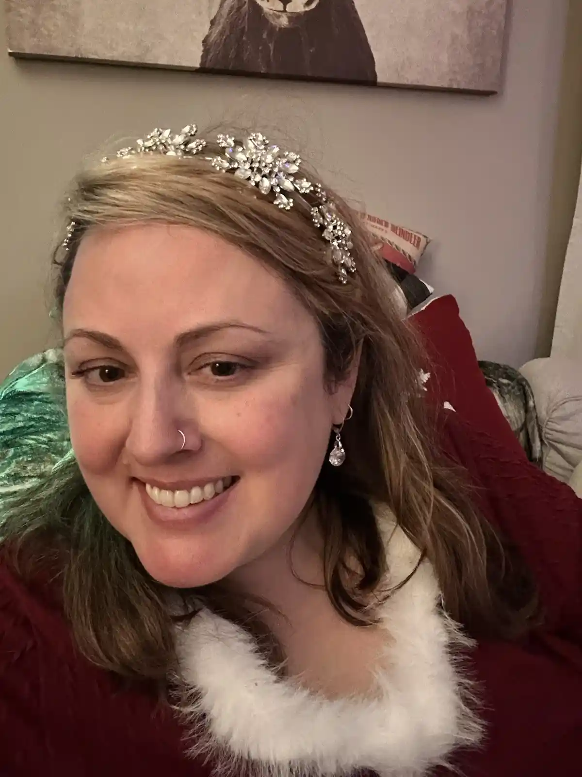 A woman with brown hair smiles while wearing a red Santa-style outfit with white faux fur trim, a jeweled snowflake…