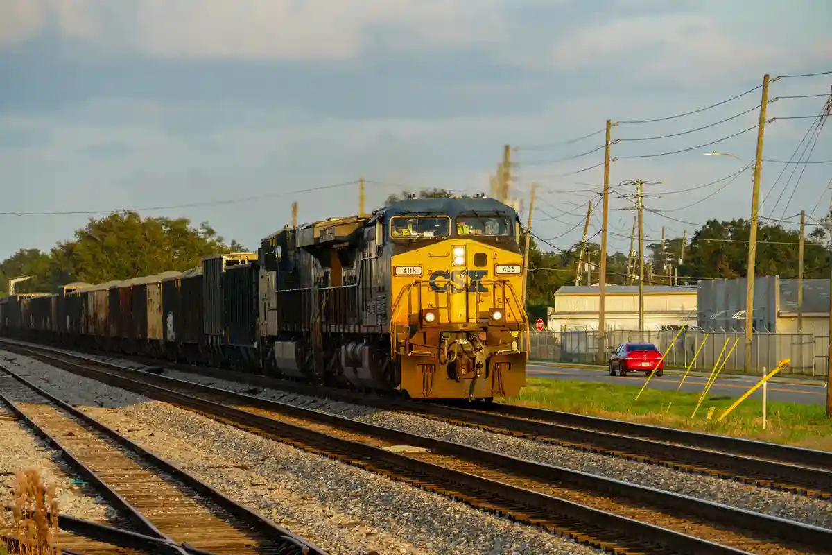 A yellow CSX freight train with locomotive number 405 is shown moving along tracks, with a red car visible on a parallel…