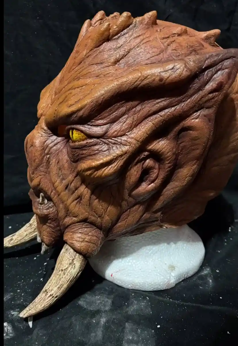 A highly detailed, brown, textured monster mask with a single glowing yellow eye, prominent tusks, and wrinkled skin sits on…