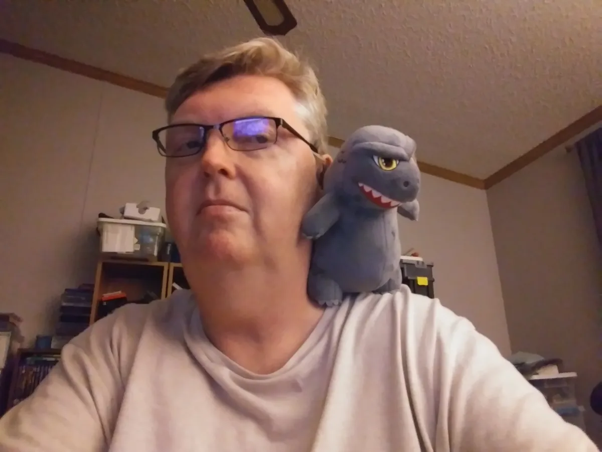 A man with glasses and a neutral expression has a small gray plush dragon with yellow eyes perched on his shoulder. The…