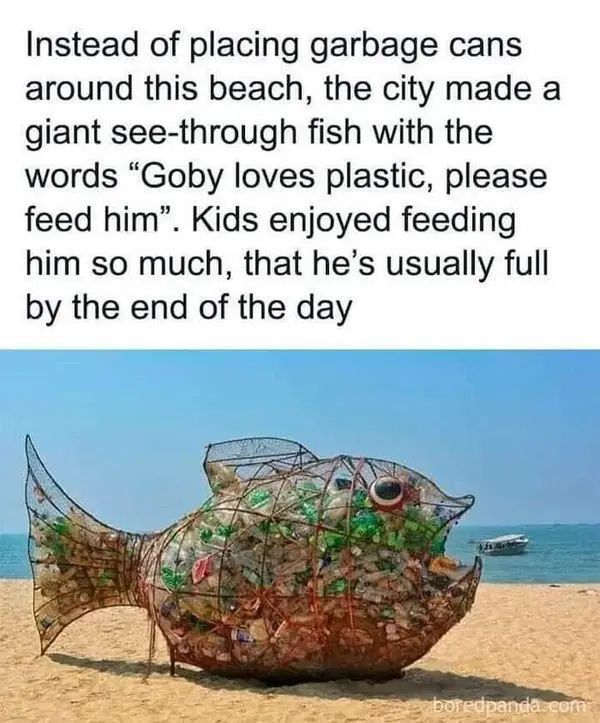 A large, transparent fish sculpture made of plastic bottles and wire stands on a sandy beach near the ocean. The fish has a…