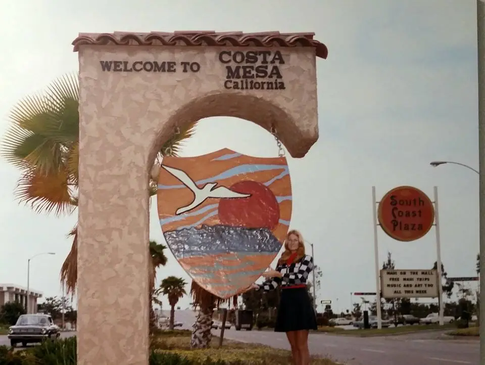 A vintage photo shows a woman standing beside a large, arched 'Welcome to Costa Mesa, California' sign featuring a painted…