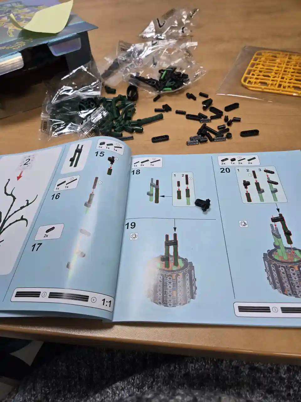 An open instruction manual for a plastic model kit lies on a wooden table, surrounded by scattered green and black plastic…