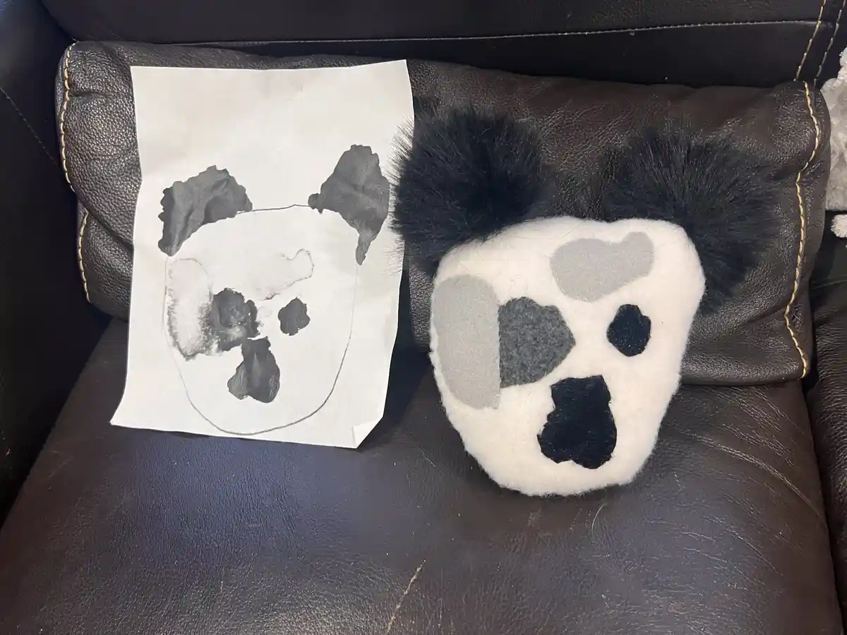 A handmade plush panda toy with fuzzy black hair and felt patches sits next to a child's crayon drawing of a panda on white…