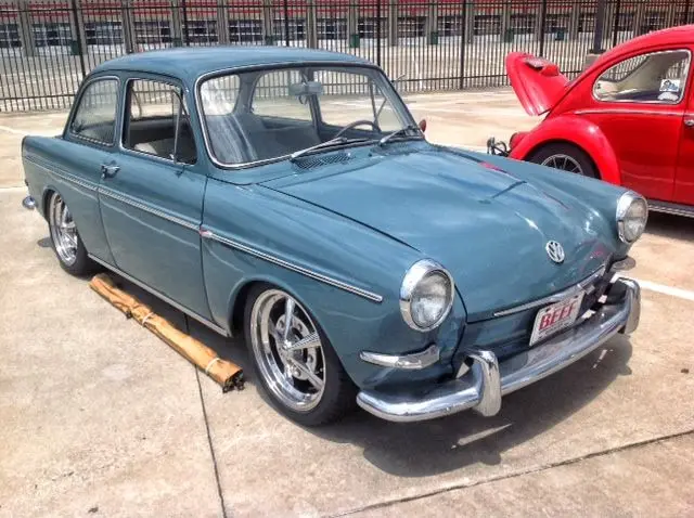 A teal vintage Volkswagen Beetle with chrome bumpers and custom wheels is parked on a concrete lot, with a wooden plank…
