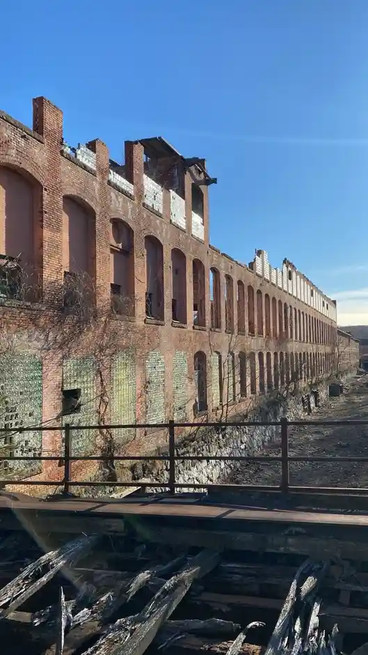 A long, dilapidated brick factory building with broken windows and missing roof sections stands under a clear blue sky…