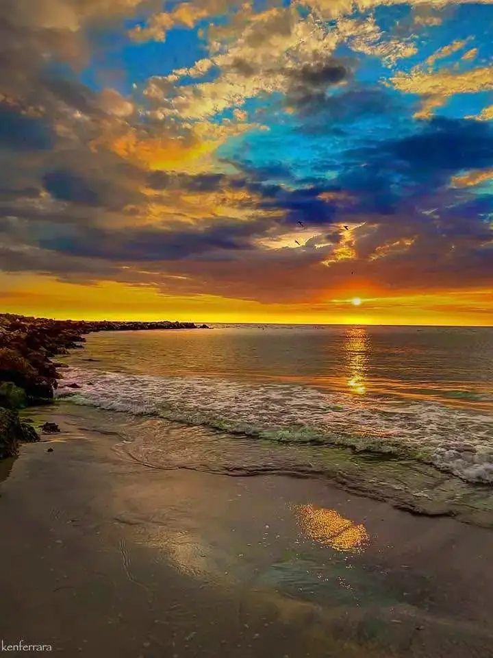 A vibrant sunset over a beach with waves lapping at the shore, clouds painted in orange, yellow, and blue, and a watermark…