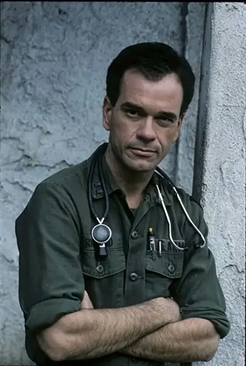 A man in a green military-style shirt with a stethoscope around his neck stands with arms crossed against a textured wall…
