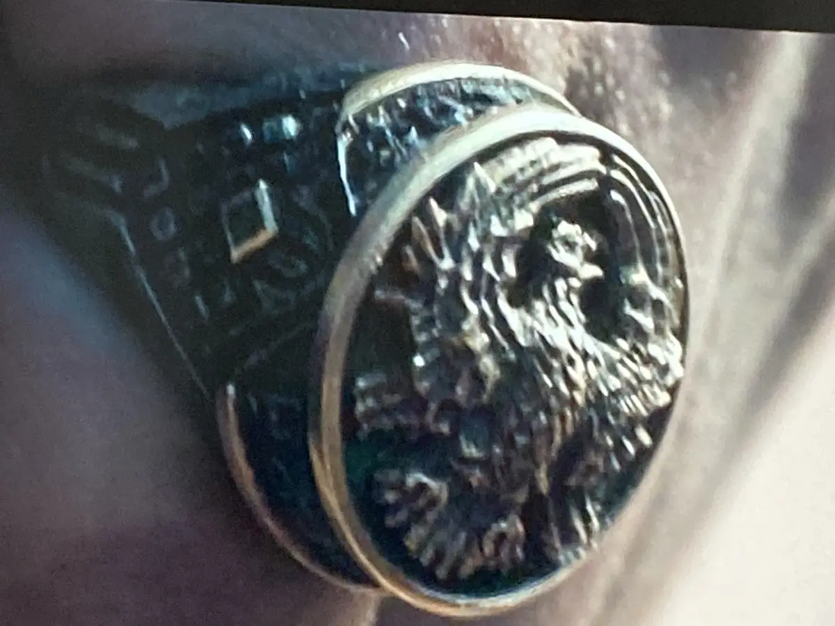 A close-up photo shows a large, ornate metal ring with a detailed, embossed eagle head on its face, worn on a finger. The…