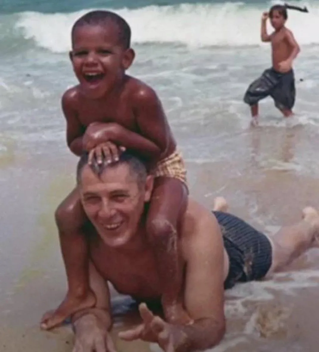 A vintage photo shows a smiling man on his hands and knees in shallow ocean water, with a young boy laughing joyfully riding…