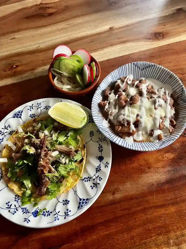 A top-down photo shows a taco on a blue-and-white floral plate next to a bowl of beans topped with white sauce, with a small…