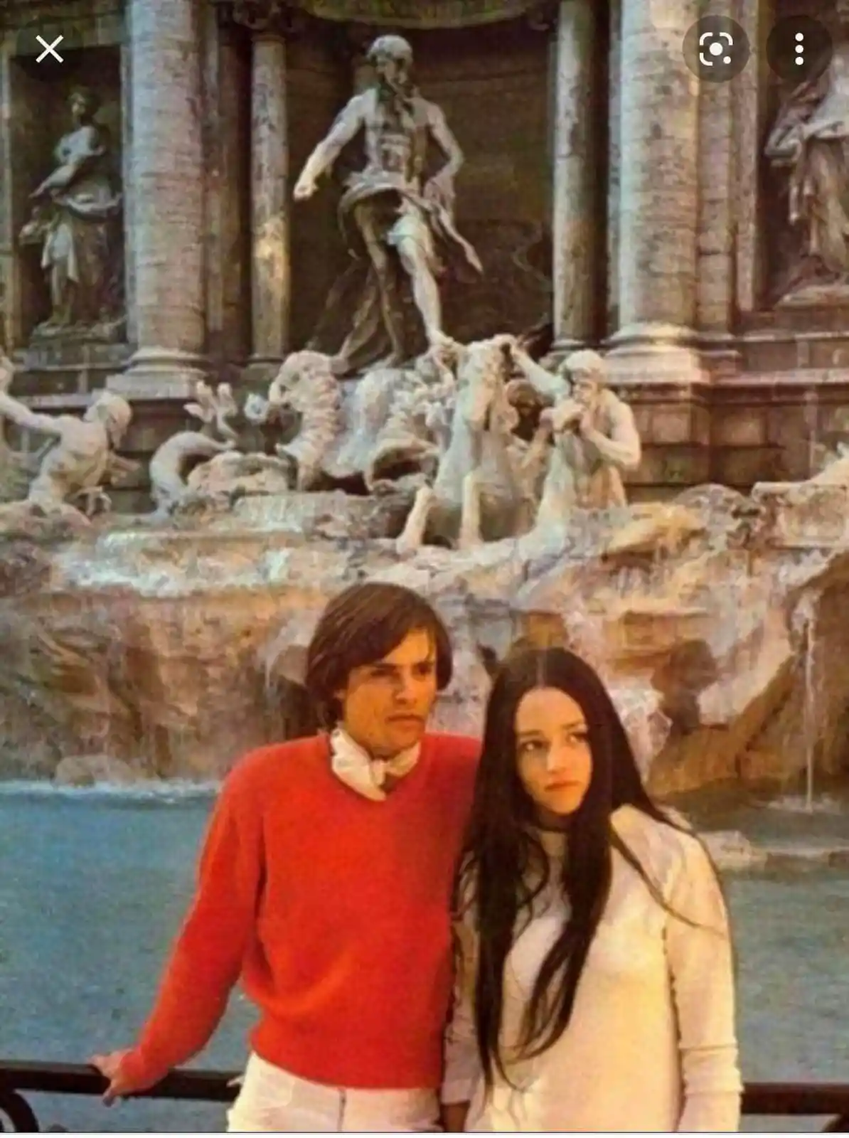 A vintage photo shows a man in a red sweater and a woman in a white top standing in front of the Trevi Fountain in Rome…