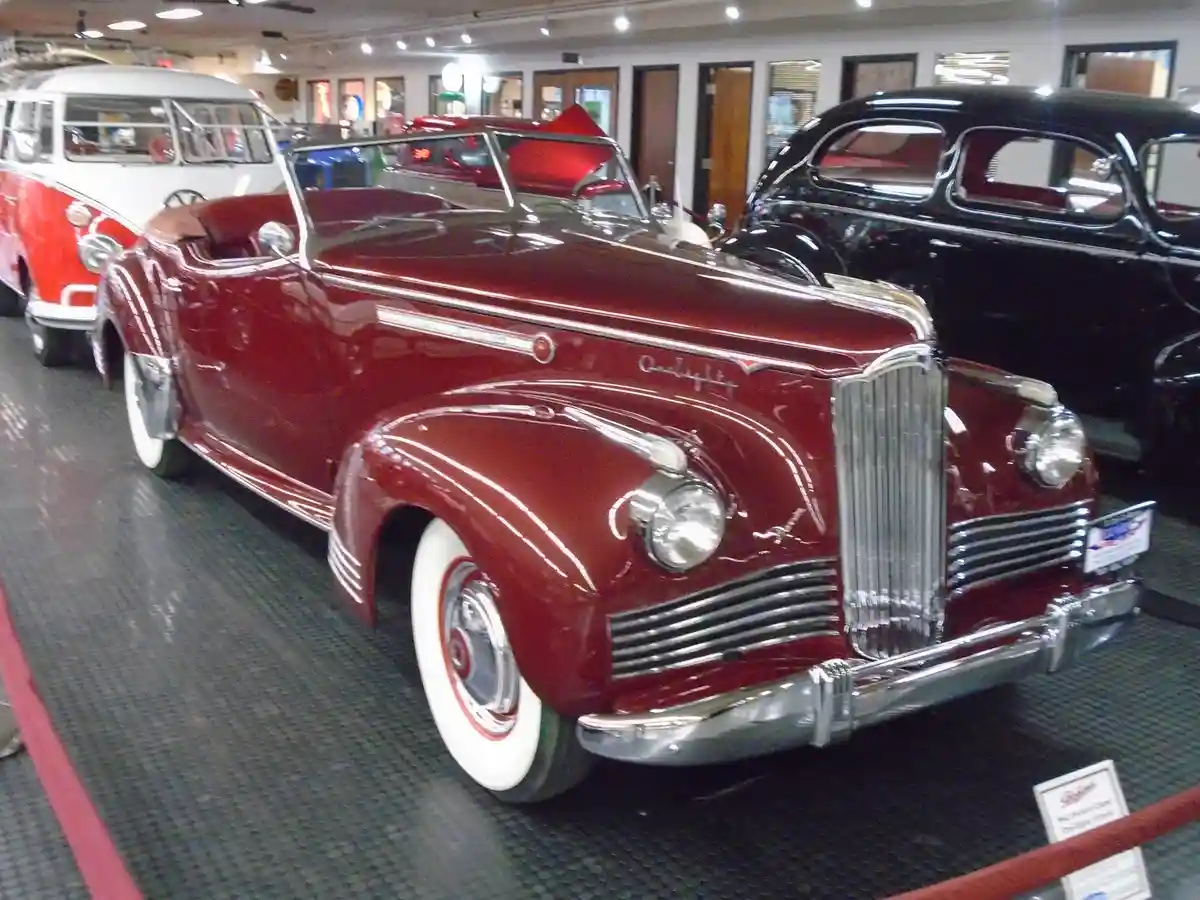 A glossy red 1940s Chrysler convertible is displayed in a museum, surrounded by other vintage cars including a red-and-white…