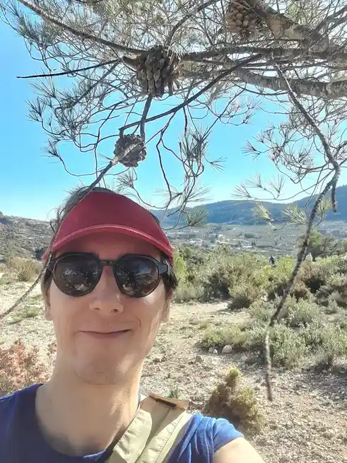 A person wearing a red cap and sunglasses takes a selfie outdoors under a pine tree, with pine cones visible in the branches…