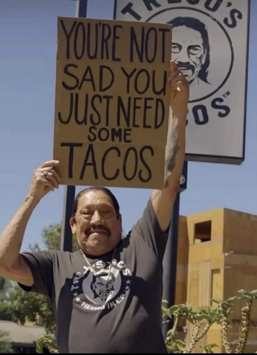 A man in a Trejo's Tacos t-shirt holds up a cardboard sign that reads 'YOU'RE NOT SAD YOU JUST NEED SOME TACOS' in front of…