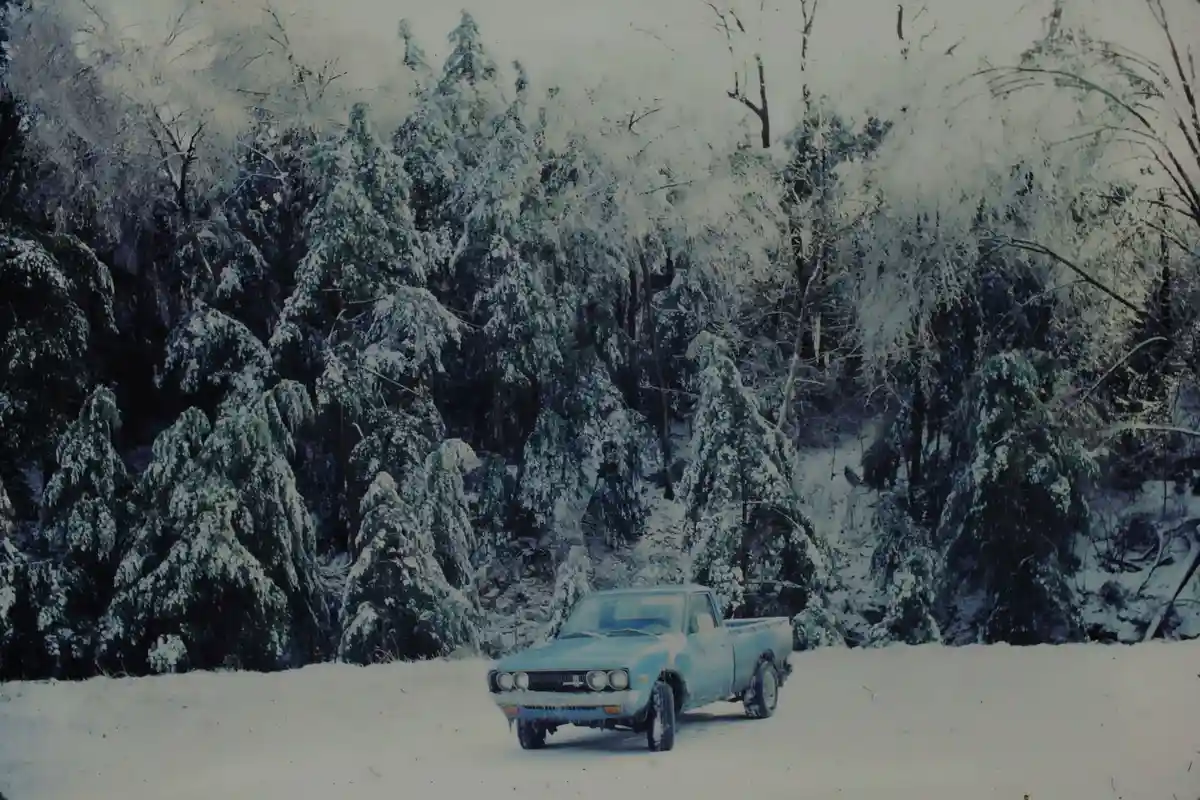 A vintage light blue pickup truck is parked in a snowy forest, surrounded by snow-covered evergreen trees. The overlaid text…