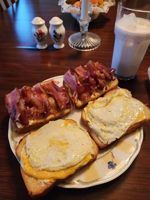 A plate of four slices of toast, two topped with fried eggs and cheese, two topped with crispy bacon, set on a wooden table…