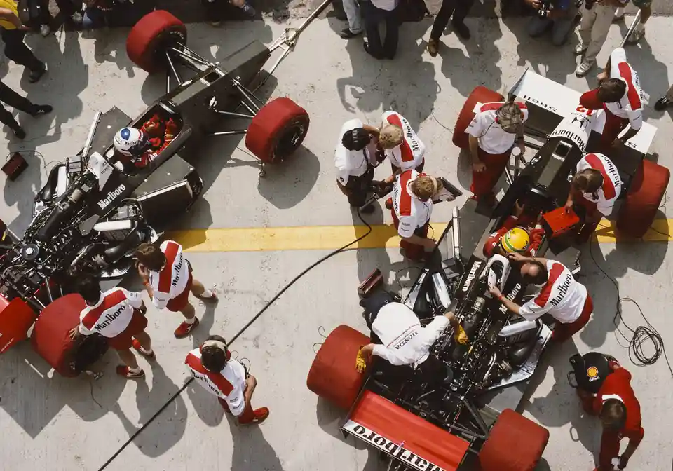An overhead photo shows two Formula 1 cars with their teams working on them; the car on the left has a driver in the…