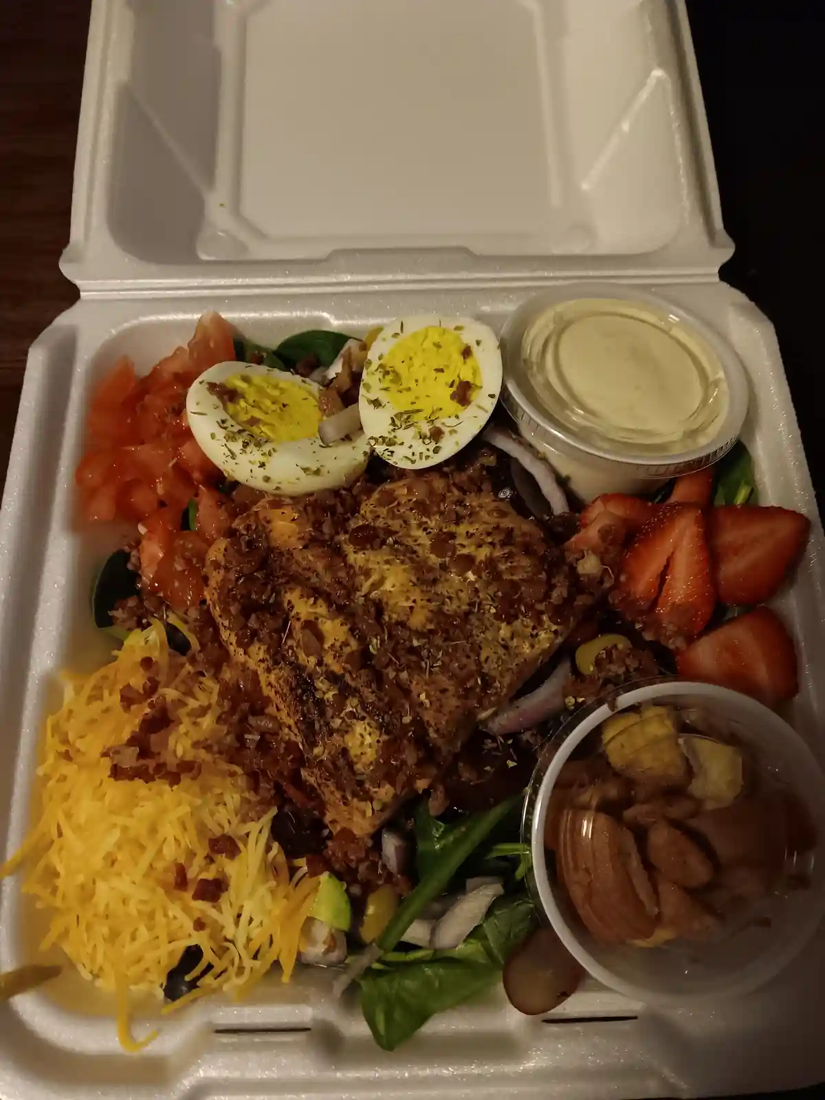 A white styrofoam takeout container holds a large, heavily loaded meal that appears to be a salad but contains ingredients…