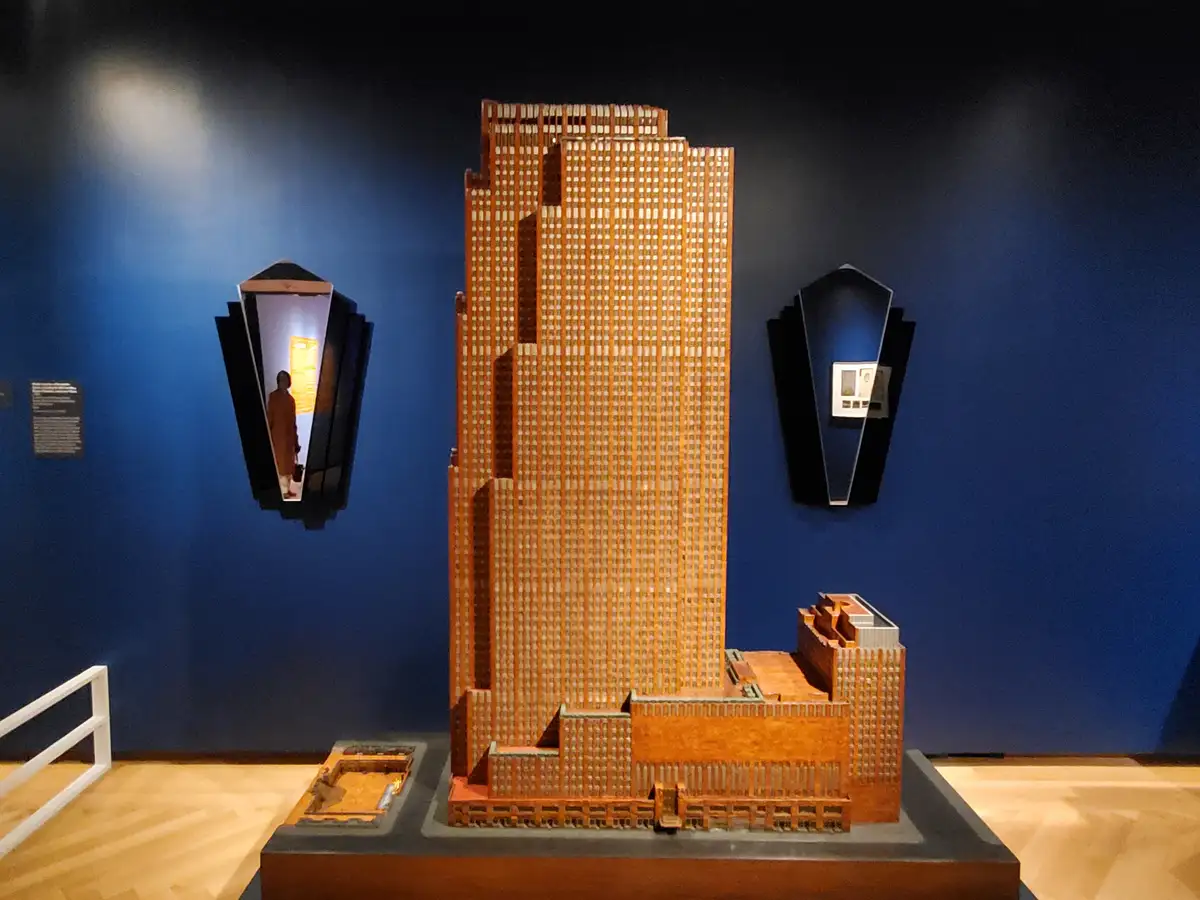 A large architectural model of a skyscraper made of orange-brown material is displayed on a pedestal in a museum with blue…