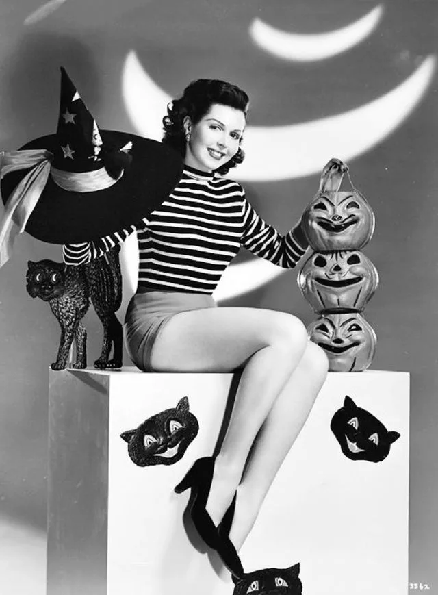 A vintage black-and-white photo shows a smiling woman in a striped shirt and skirt sitting on a white block, holding a…