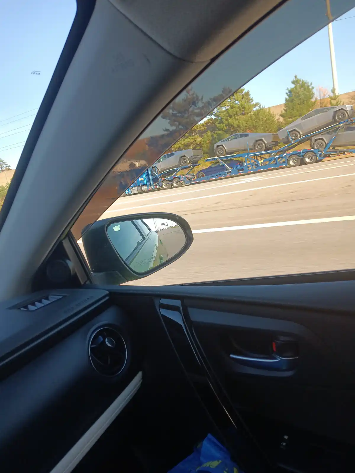 A photo taken from inside a car shows a blue car carrier truck loaded with multiple silver Tesla Cybertrucks on a highway…