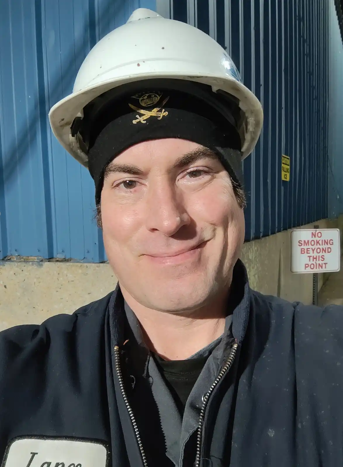 A man wearing a white hard hat over a black beanie with a pirate skull and crossbones emblem smiles at the camera while…