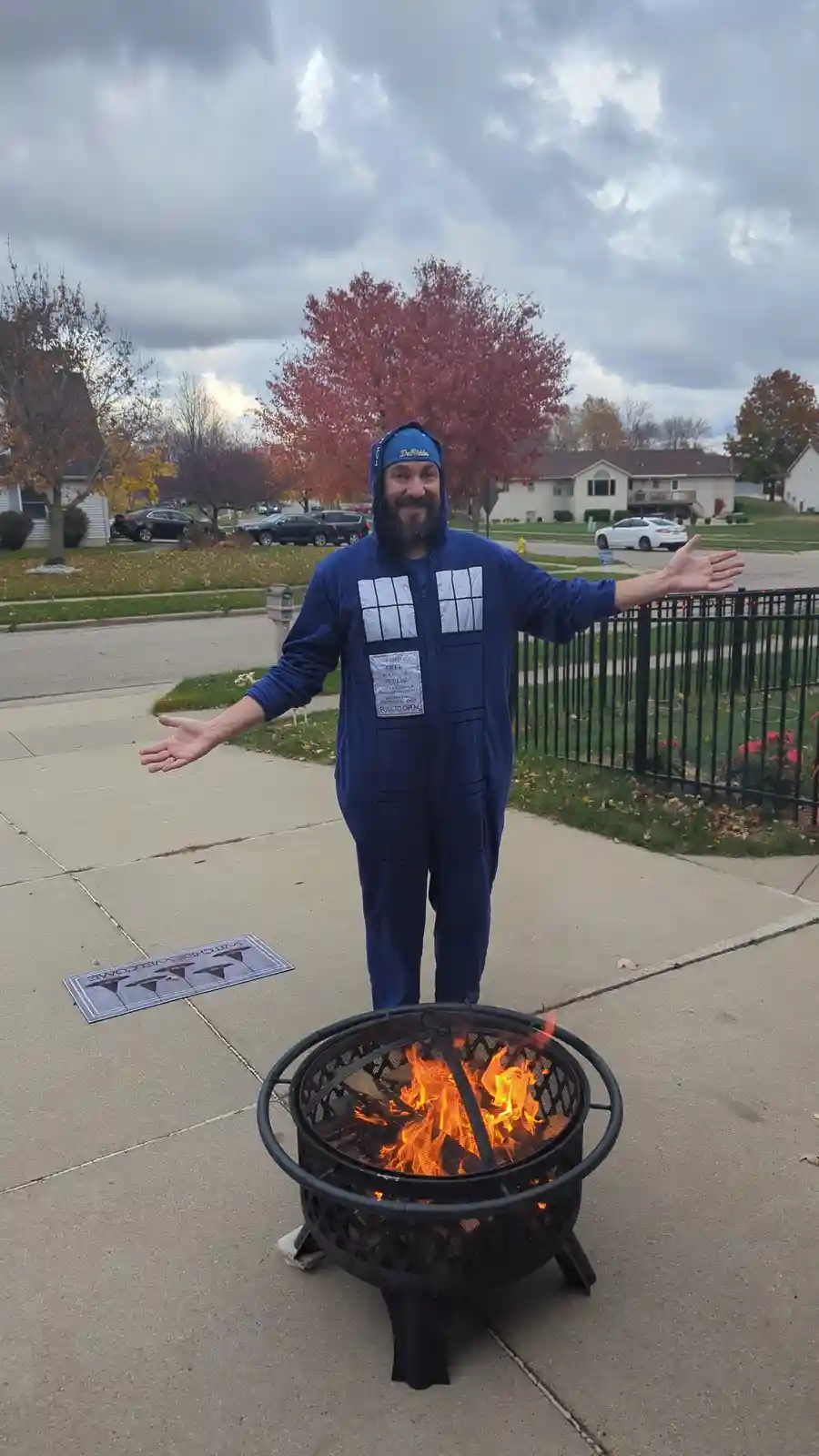 A man in a blue onesie resembling the TARDIS from Doctor Who stands with arms outstretched on a sidewalk next to a lit fire…