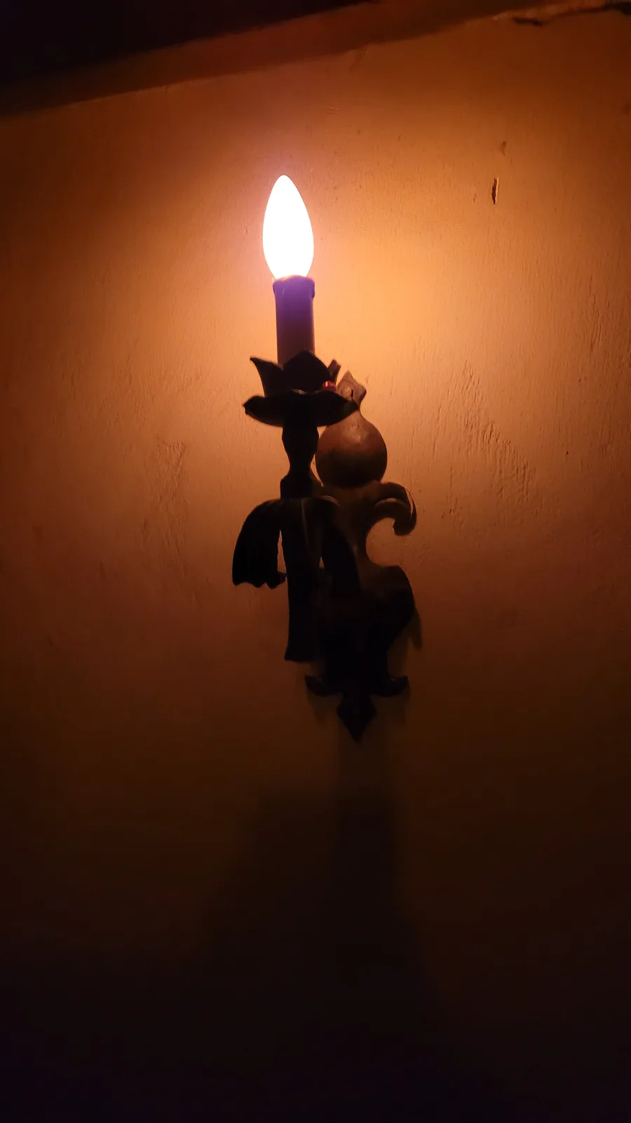 A close-up of an ornate, dark metal wall sconce holding a lit bulb that is shaped and positioned to resemble a candle. The…