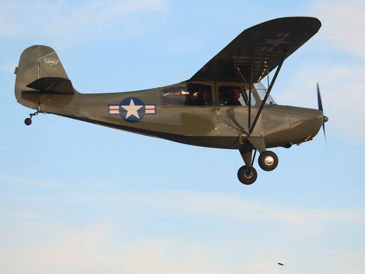 A vintage olive-drab biplane with U.S. military markings, including a star insignia on the fuselage and wing, is flying low…