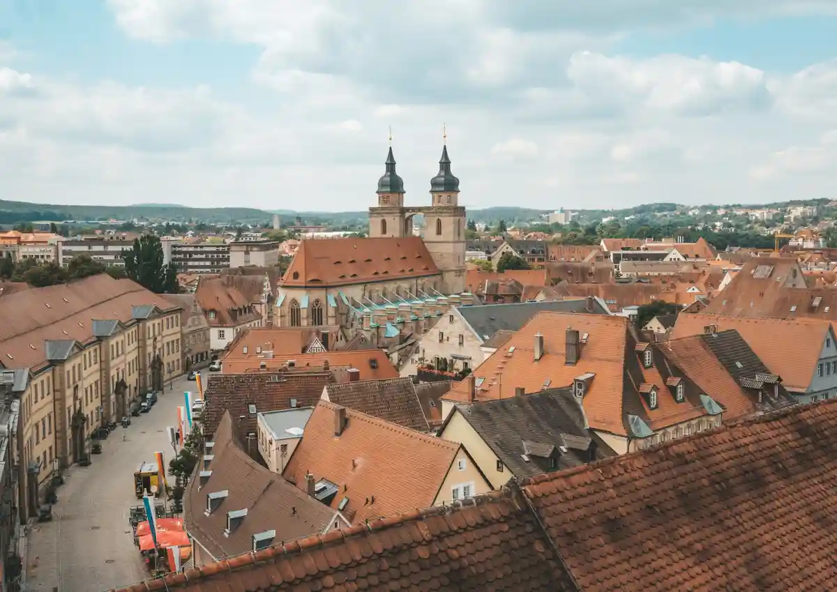 A scenic aerial view of a European city with red-tiled roofs, a prominent church with two dark spires, and distant hills…