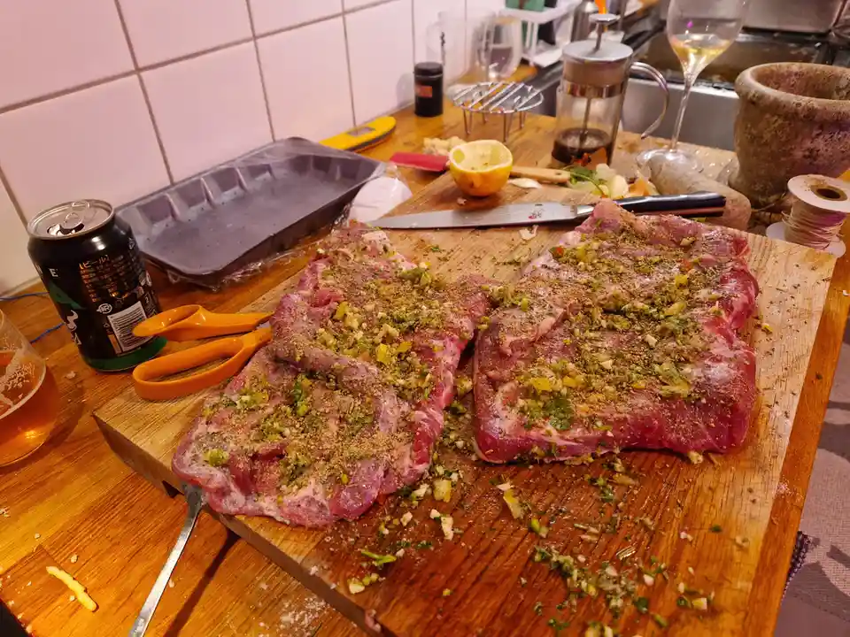 Two large raw meat cuts, likely ribs, are heavily seasoned with herbs and spices on a wooden cutting board in a kitchen…